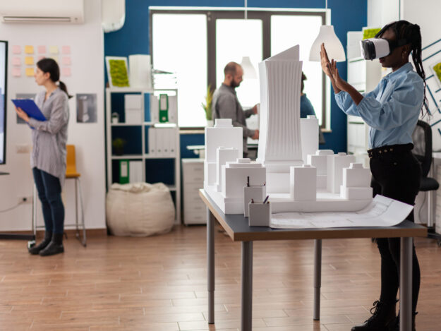 Architect standing in modern architectural office using virtual reality goggles Architect standing in modern architectural office using virtual reality goggles to view 3d plan of white foam maquette. Engineer working with vr headest next to table with model of urban project.