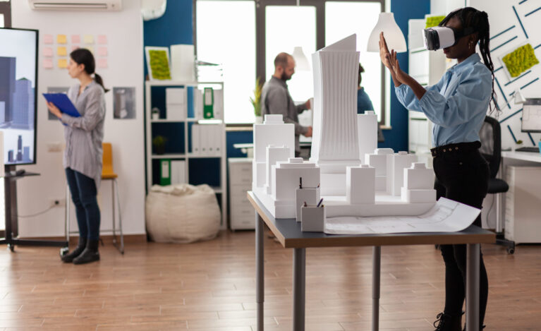 Architect standing in modern architectural office using virtual reality goggles to view 3d plan of white foam maquette. Engineer working with vr headest next to table with model of urban project.